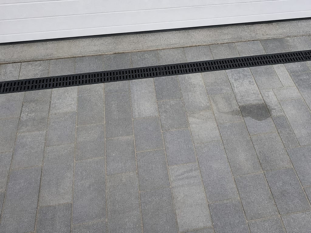 Black channel drain set across block paving in front of a garage door